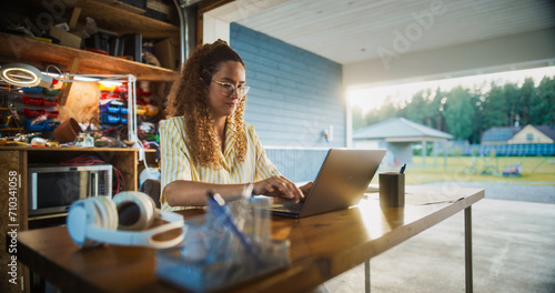 Happy Hispanic Woman Using Laptop Computer In Opened Garage. Successful Female Digital Entrepreneur Running An E-commerce Startup From Home, Checking Emails, Filling Online Orders, Researching Market.