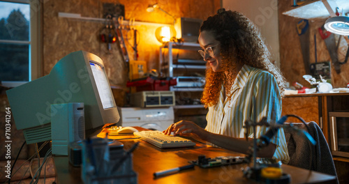 Hispanic Female Engineer Using Old Desktop Computer In Nineties Retro Garage In The Evening. Intelligent Woman Writing Code, Working On Innovative Portable Device, Starting Tech Startup Company.