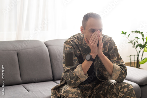 Thoughtful military man staring aside, holding palms by mouth, sitting on couch at home. Young soldier visiting psychologist, suffering from posttraumatic stress, closeup photo, copy space