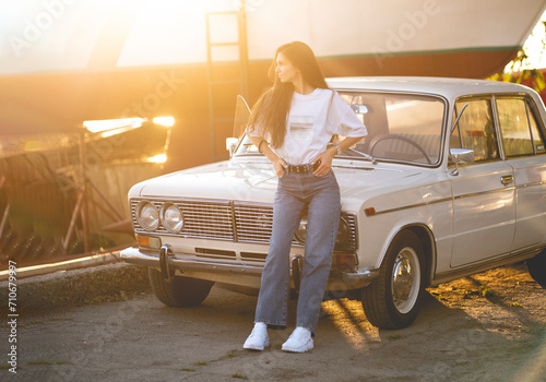 young girl in casual attire stands beside a classic retro car in warm glow of a sunset sun