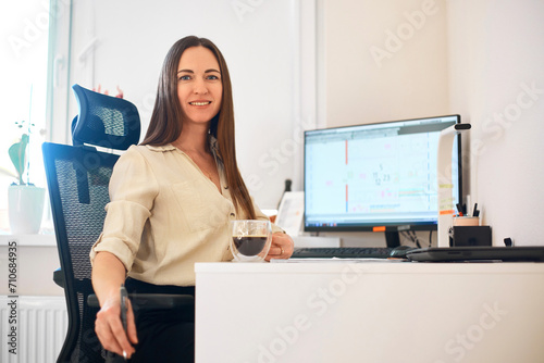 Portrait of a happy female designer estimator sitting at the table with a notebook and calculator, counting the balance and typing on a laptop, finishing her work with a cup of coffee.