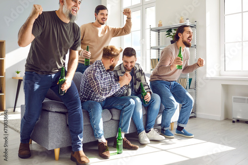 Group of football fans watching sport match on TV together. Emotional male friends sitting on sofa drinking beer and celebrating victory of their team at home. Friendship, entertainment concept
