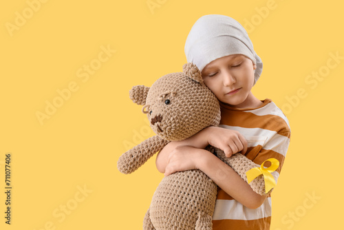Cute little boy after chemotherapy with yellow ribbon and teddy bear on color background. Childhood cancer awareness concept