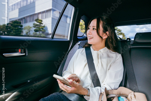 スマホを持ちながら自動車の後部座席で窓の外を眺めるビジネスウーマン（出張・検索・アプリ） 