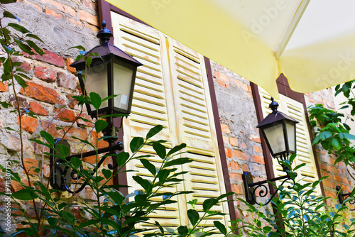 Old windows in wooden frames and with closed shutters, with green plants around, metal street lanterns and an old brick wall. Part of the interior of an outdoor cafe. Torun, Poland, August 2023