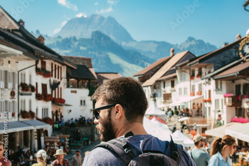 Young caucasian man tourist with beard and wearing sunglasses traveling with backpack exploring the european city Gruyères in Fribourg, Switzerland on summer season. SHOTLISTtravel.