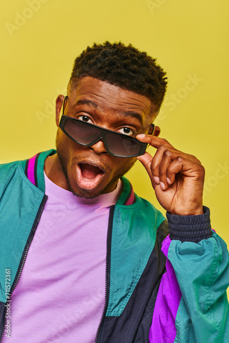 impressed african american man with open mouth holding sunglasses and looking at camera on yellow