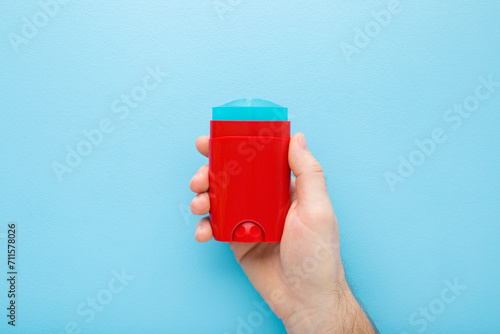 Young adult man hand holding and showing red plastic stick of armpit deodorant on light blue table background. Pastel color. Male daily beauty product. Closeup. Top down view.