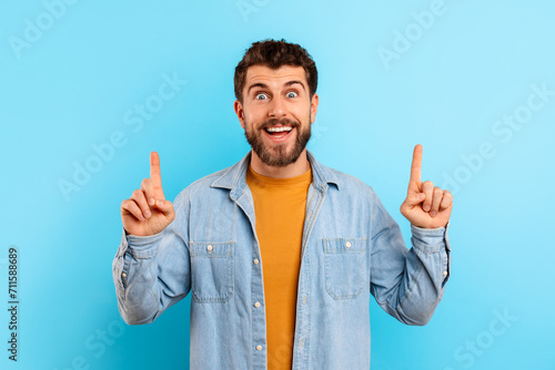 Excited young bearded man pointing fingers up on blue background