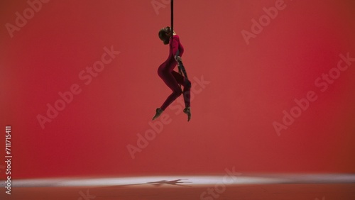 Female gymnast isolated on red studio background. Girl aerial dancer in bodysuit on acrobatic trapeze with straps perform element.