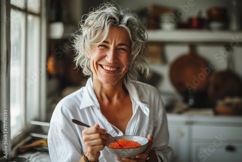 Smiling beautiful elderly woman eating red caviar by the spoon in the kitchen. Concepts: healthy lifestyle, active longevity, proper nutrition, healthy fats, healthy skin