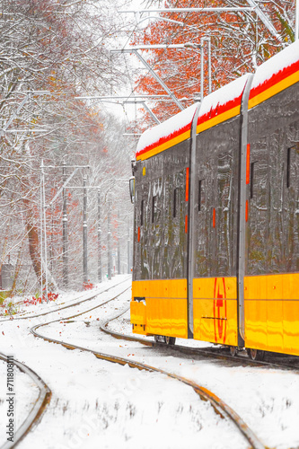 Jadący tramwaj w Warszawie na Bielanach w scenerii zimowej przy padającym śniegu pośród drzew..