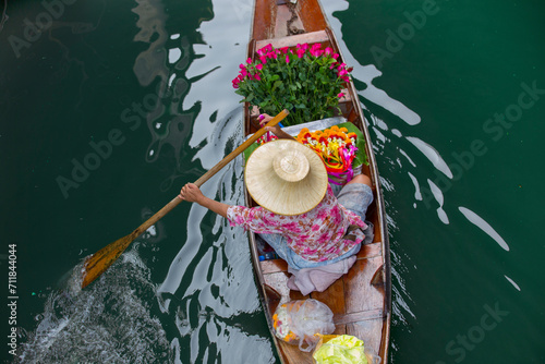 boater in Thailand shot from above, she is carrying flowers to the market and wearing a bamboo hat and holding an oar at the floating market Damnoen saduak near bangkok