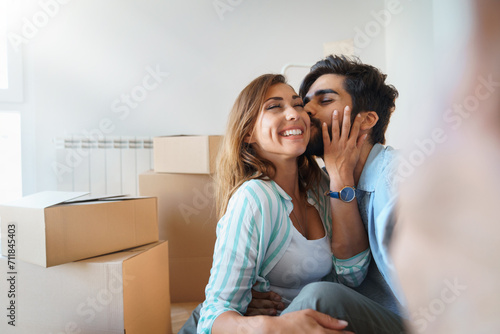 Loving young married couple hugging and kissing while taking selfie during relocation, surrounded with cardboard boxes around.