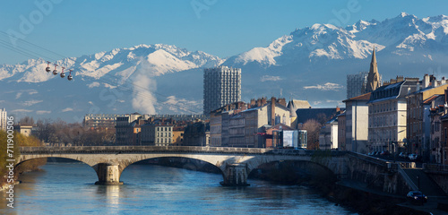 Cityscape of Grenoble with famous cable car against backdrop of snowy Alps in sunny day, France
