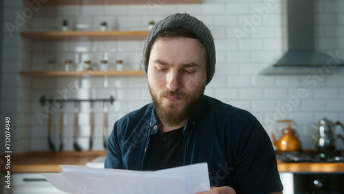 Portrait of young adult man with beanie sitting in modern kitchen at home, talking, looking the reports in video conference call. Confident male have video conference, remote chat