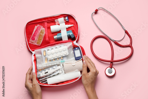 Female hands with first aid kit and pills on pink background