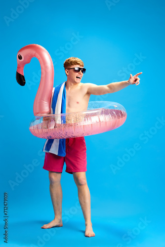 Young cheerful relaxed guy in swimming circle enjoying summer trip, having fun against blue studio background. Concept of emotions, youth, leisure time, summer vacation