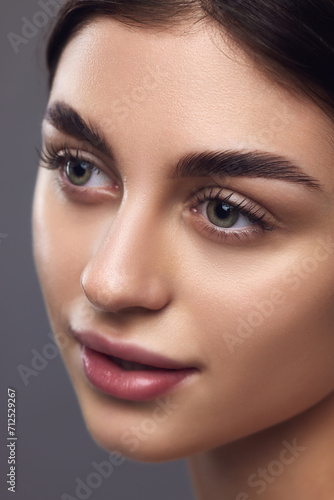Close up portrait of female model face with with perfect lips and eye makeup posing against grey studio background. Concept of eyebrow styling product, natural beauty, full brows.