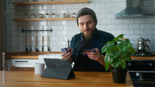 Anxious man with beanie sitting in kitchen at home takes his credit cards out of his pocket and tries them one by one to find the credit card that has not reached its limit