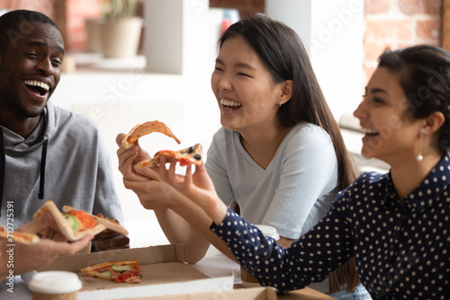 Smiling multiethnic young people have fun eating tasty Italian fast food from takeaway delivery, overjoyed multiracial students laugh joke enjoying delicious pizza on lunch break in class together