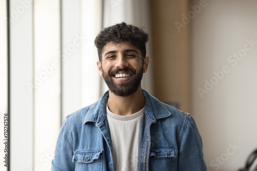 Positive handsome Arab entrepreneur man in casual clothes standing for shooting indoors, looking at camera with toothy smile, laughing. Cheerful young bearded project manager guy head shot portrait