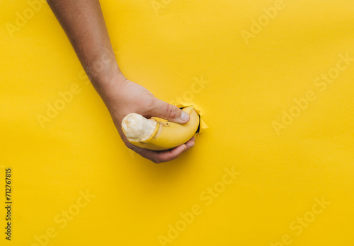 A man's hand holds a penis-shaped banana from a torn hole in yellow paper. Comic concept of masturbation, erection and arousal.
