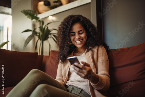 Happy young Latin woman sitting on sofa holding mobile phone using cellphone technology doing ecommerce shopping, buying online, texting messages.