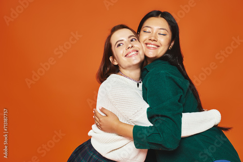 happy interracial friends in casual clothing embracing and smiling on orange background, women