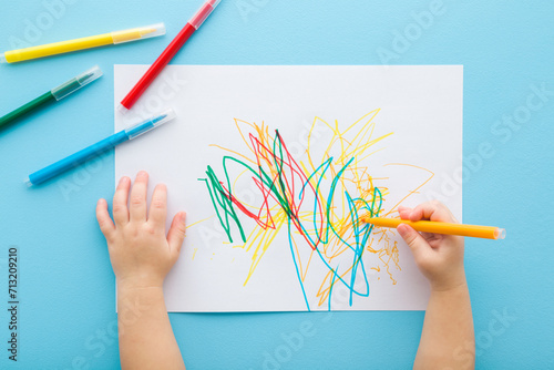 Baby hand holding marker and drawing colorful scratches lines on white paper. Blue table background. Pastel color. Closeup. Toddler development. Learning painting. Point of view shot. Top down view.