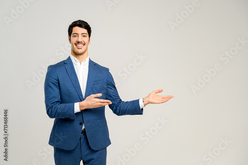 Young male business professional in a stylish blue suit pointing aside, his open hand suggesting a presentation or a welcoming introduction, set against a plain background