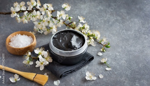 Preparing black clay cosmetic mask with almond spring flower decoration on gray background with copy space
