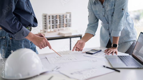 Two engineer architect pointing on blueprint of sketching interior architectural building to discussion technical for construction plan and building model while working together in workplace site