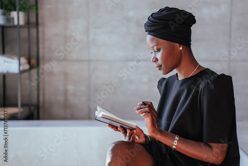 Contemplative African woman reading book, black attire, headwrap, modern space
