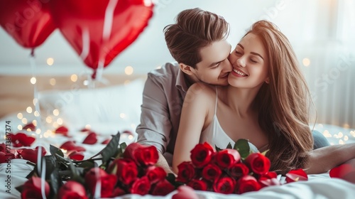 Beautiful young couple at home. Hugging, kissing and enjoying spending time together while celebrating Saint Valentine's Day with red roses on bed and air balloons in shape of heart on the background.