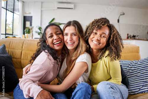 Portrait of three attractive female room mates looking smiling at camera. Group of cute cheerful girls hugging sitting on couch at home. Happy friends posing for photo indoors. Generation z and rent.
