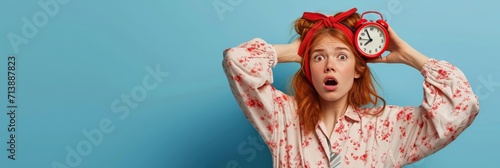 Anxious, panicking young redhead girl missed interview, overslept, holding red alarm clock and touching head frustrated with concerned face, wearing nightwear and sleep mask, blue background. 