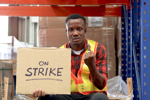 Angry unhappy African worker man wearing safety vest and giving the middle finger with strike banner placard sign at cargo logistic warehouse. Striking worker protesting at workplace.