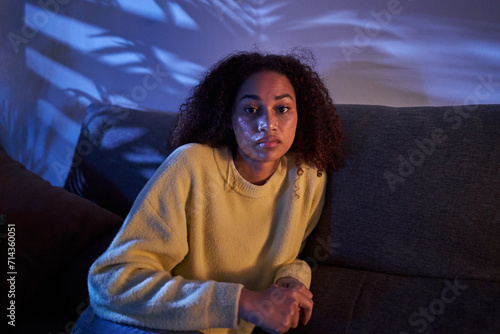 Portrait young African American woman looking serious on camera sitting alone on sofa at night home. Afro female posing with sad expression indoors. Photo of lonely, worried and dissociated black girl