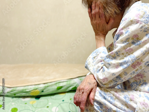 畳んだ布団の前で頬杖をつく寝間着姿の高齢女性