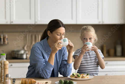 Beautiful young mom and little preschool kid having breakfast at home, sitting at kitchen table, drinking hot beverage, eating sandwiches with fresh vegetables. Mother feeding child