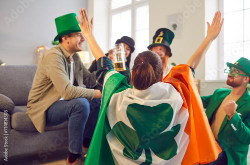 Happy smiling friends celebrating Saint Patrick's Day at home sitting in a circle in green clothes and hats. Young men and women talking and having fun on party drinking beer with a flag of Ireland
