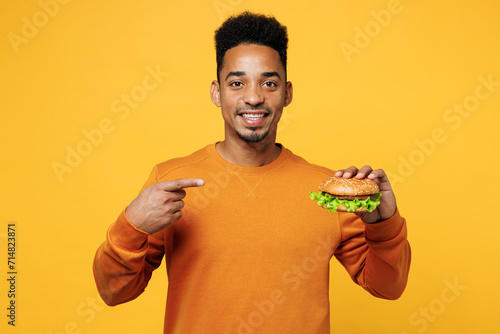 Young man wear orange sweatshirt casual clothes point index finger on burger isolated on plain yellow background studio portrait. Lifestyle concept. Proper nutrition healthy fast food choice concept.