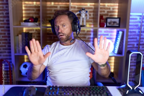 Middle age man with beard playing video games wearing headphones moving away hands palms showing refusal and denial with afraid and disgusting expression. stop and forbidden.