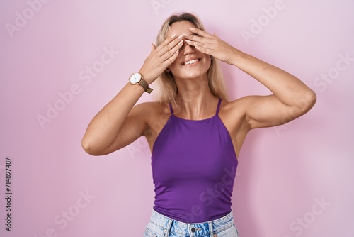 Young blonde woman standing over pink background covering eyes with hands smiling cheerful and funny. blind concept.
