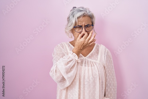 Middle age woman with grey hair standing over pink background smelling something stinky and disgusting, intolerable smell, holding breath with fingers on nose. bad smell