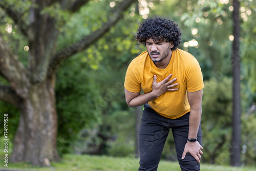 Tired young Indian male athlete standing bent over in park and holding hand to chest, feeling severe pain and shortness of breath after jogging