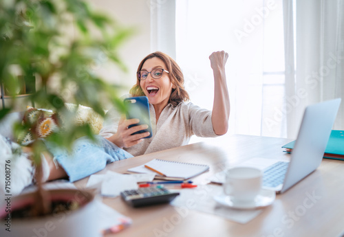 Beautiful middle-aged woman in glasses joyfully laughing at smartphone and making YES gesture at home office. Small business, successfully investments, bull market or money savings concept image.