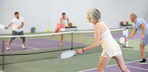 Focused active aged woman playing friendly pickleball match on indoor court, rear view. Senior people sports concept..