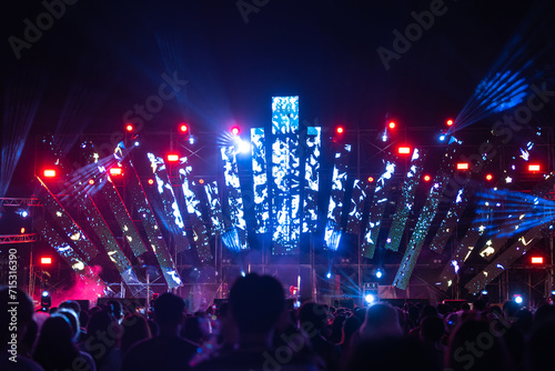 In midst of night a concert festival main event draws an enthusiastic unrecognizable crowd all cheering in front of brilliantly illuminated stage. lens flare adds to fun during this music festival.